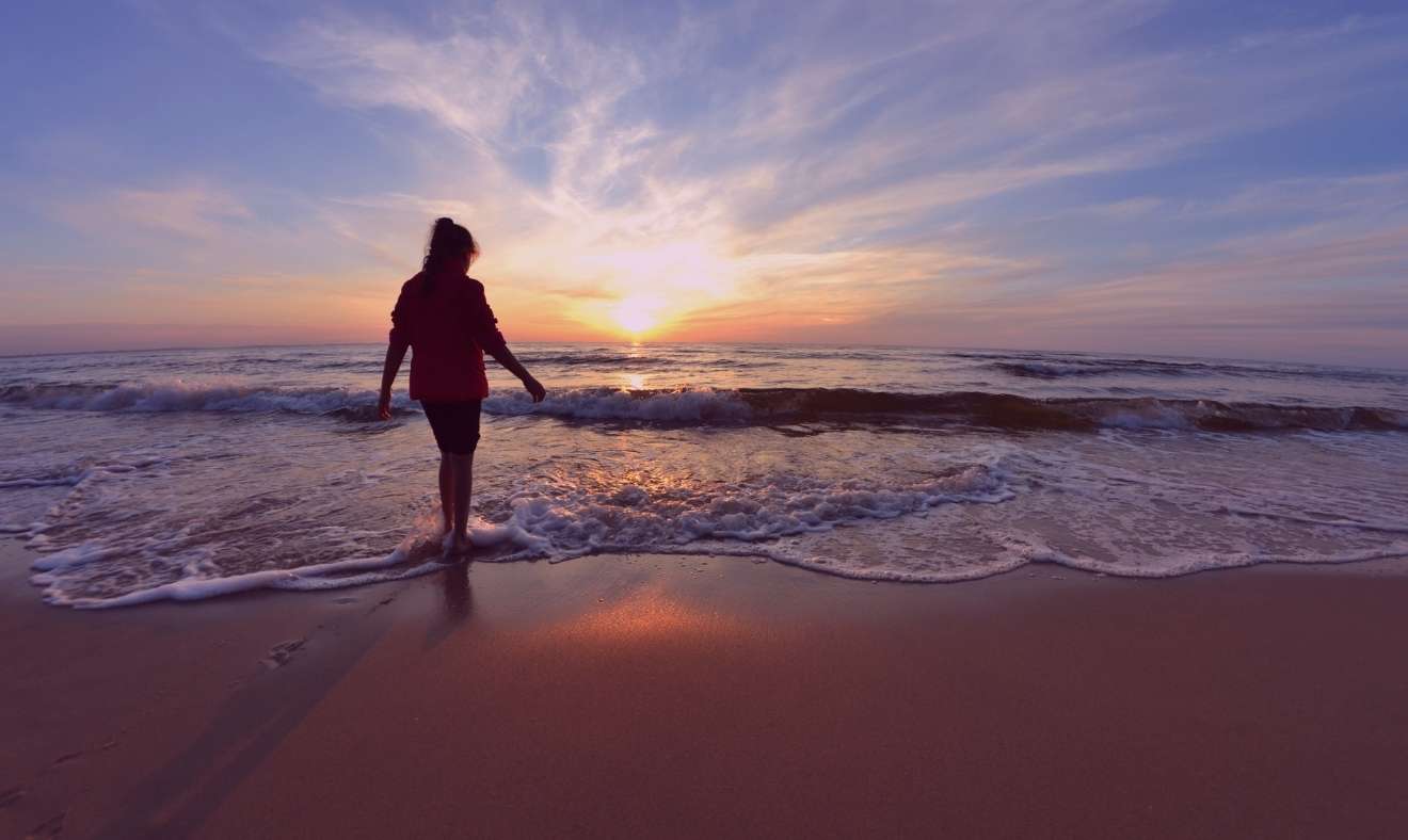 Vrouw loopt op het strand, de zon daalt neer op de horizon
